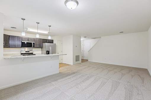 A spacious, empty room with a carpeted floor and a kitchen area in the background.