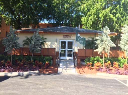 A building with a brown roof and a white door is surrounded by green plants.