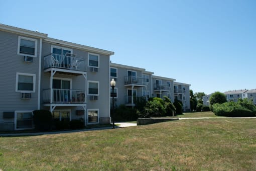 a row of grey apartment buildings with balconies and a grassy area in front of them