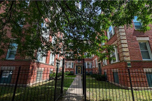A black metal gate leads to a brick building with a green lawn.