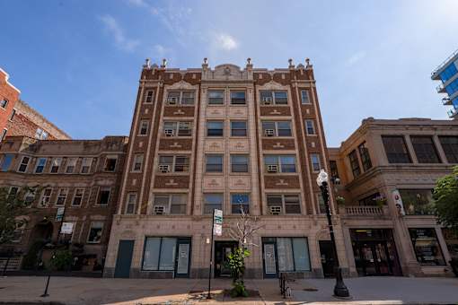A large brown building with many windows is in the middle of a city street.