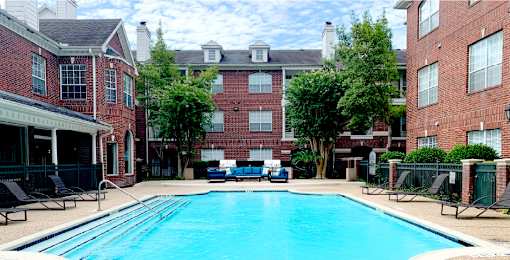 Luxury apartments with red brick exteriors, large swimming pool with spacious sundeck, grill, beautiful landscaping, lush landscaping, chaste trees, and pool-side lounge seating at Tuscany Apartments in Houston.