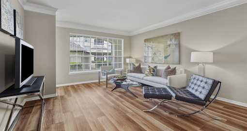 Spacious living room with wood laminate flooring, oversized window, gray painted walls, and decorative crown molding at Village on the Parkway Apartments in Houston Texas