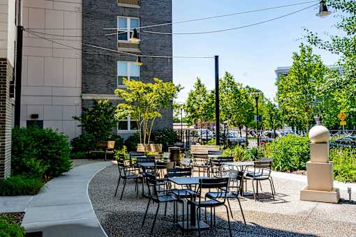 A patio with tables and chairs is surrounded by greenery and a building.