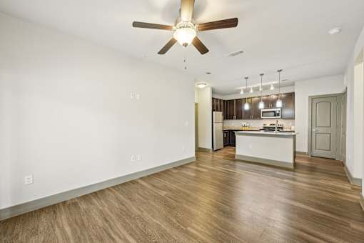 A large open room with a ceiling fan and wooden floors. The kitchen with unique lighting fixtures can be seen in the background.