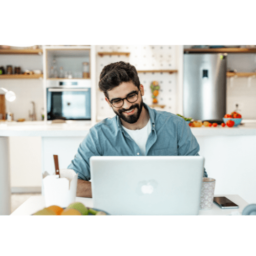 A man with glasses is working on a laptop in a kitchen.
