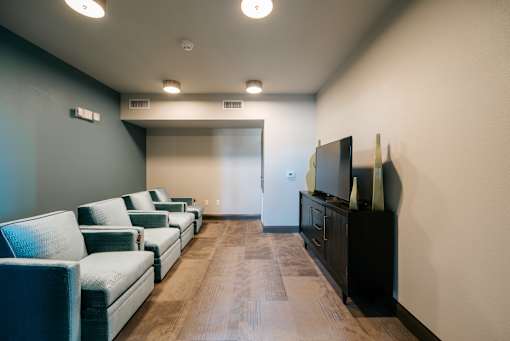 A row of grey couches are lined up in a room with a wooden floor and a wall with a television on it.