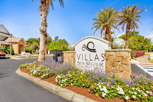A sign for Villas Bon Secour sits in front of a flower bed.