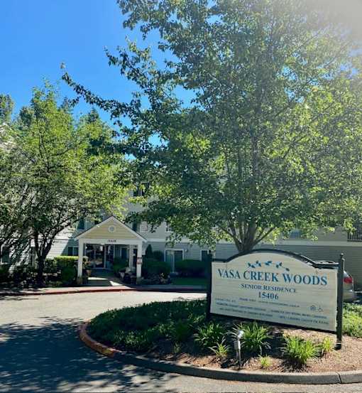 A sign for Vasa Creek Woods Senior Residence is in the foreground of a tree-lined street.