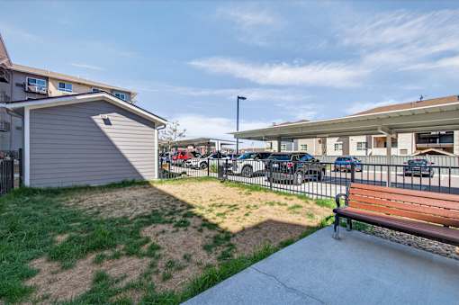 A bench is situated on a concrete area with a grassy area and a building in the background.