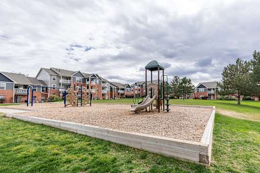 A playground with a sandbox and a slide in the foreground and apartment buildings in the background.