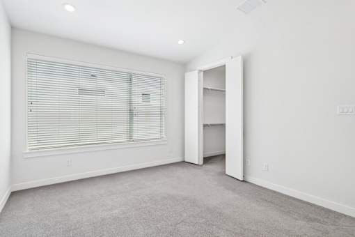 A room with a carpeted floor, a window with blinds, and a door leading to another room.