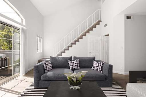 A grey couch with pillows is in the middle of a room with a staircase.
