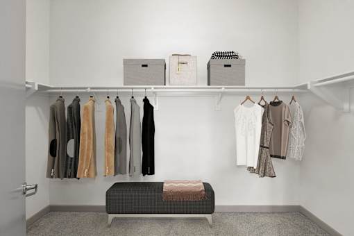 A white closet with clothes hanging and a bench under the clothes.