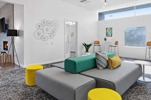 A living room with a grey sofa, yellow ottoman, and a white wall with a black and white drawing.