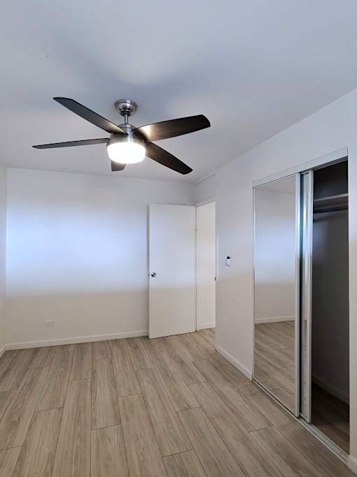 A room with a ceiling fan and light fixture, hardwood floors, and white walls with a door on the right.