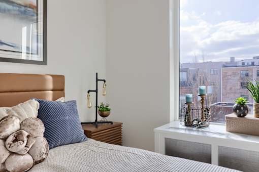 A bedroom with a bed, a nightstand with a plant, and a window overlooking a cityscape.