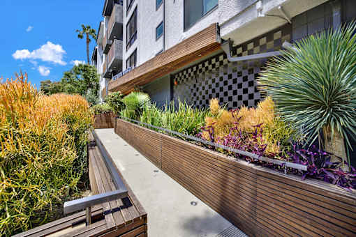 A wooden bench sits on a concrete path next to a wooden fence with a variety of plants. at Westwood Riviera Apartments, Apartments, Los Angeles, CA