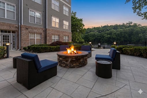 A patio with a fire pit surrounded by chairs and a building in the background.