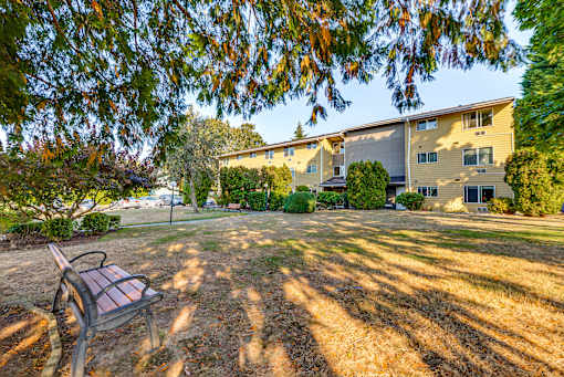 A bench is in the foreground of a grassy area with apartment buildings in the background.
