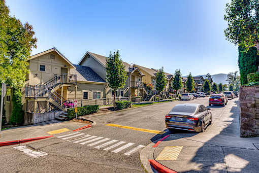 A street view of a residential area with houses on the left and cars driving on the road.