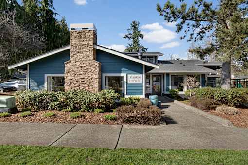 A blue building with a stone chimney and a sign that reads "LEASING OFFICE" is surrounded by greenery and a sidewalk.