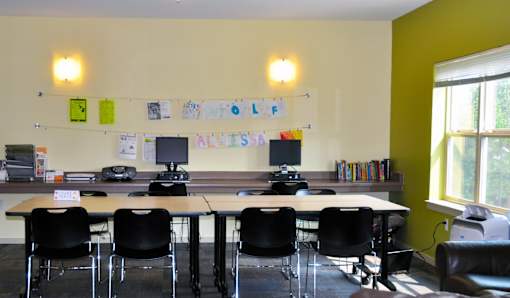 A room with a long table and chairs, a computer desk, and a bulletin board with the word "ALL'S" on it.