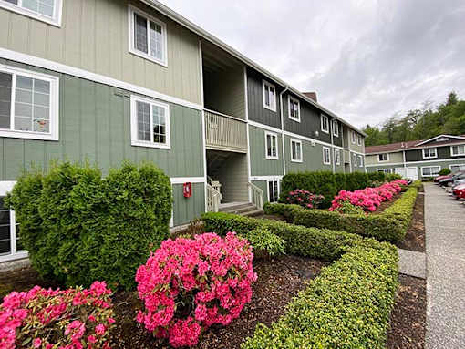 A building with a green and white exterior has a balcony and is surrounded by bushes and flowers.
