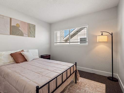 A bedroom with a bed, pillows, a nightstand, a lamp, and a window with blinds.