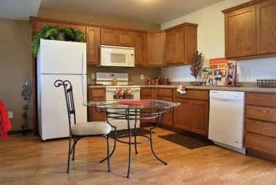 A kitchen with a white fridge, a glass table and a chair, and wooden cabinets.