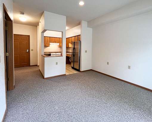 A room with a carpeted floor and a kitchen area in the background.