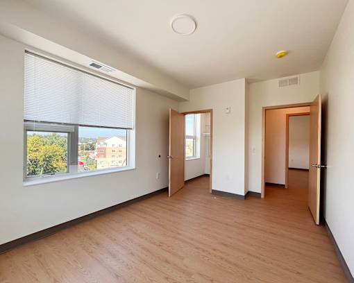 A room with wooden floors and white walls, a window with blinds, and a door on the right.