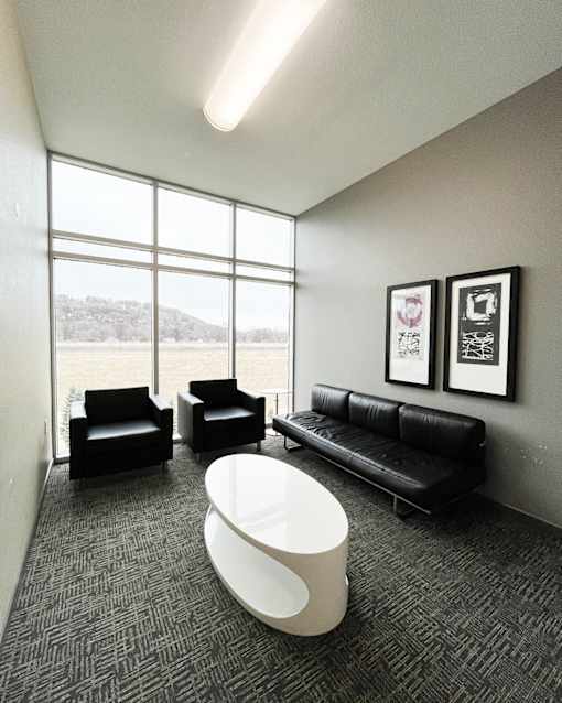 A white table is in the middle of a grey carpeted room with two black chairs and a black couch.