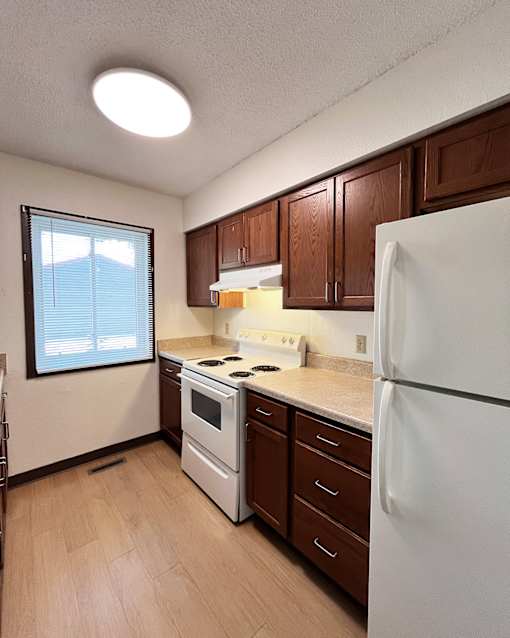 A kitchen with a white refrigerator, brown cabinets, and a window with blinds.