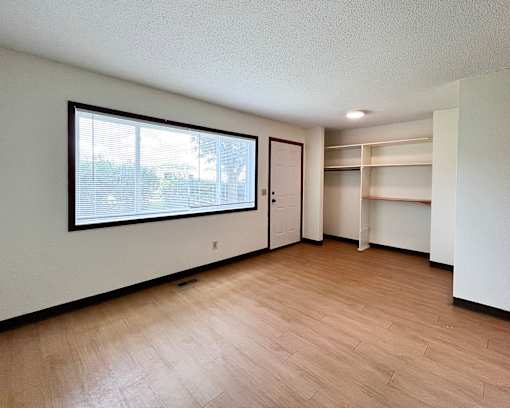 A room with a window, wooden floors, and a white wall with a door and shelves.