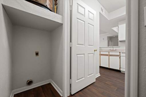 Small laundry closet with washer and dryer hookups, including electrical outlet, water connections, and vent opening, located near the kitchen in a Crescent Apartments unit.
