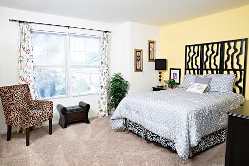 A bedroom with a bed, chair, ottoman, and window.