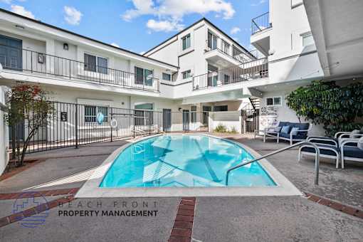 A pool surrounded by a patio with chairs and a building in the background.