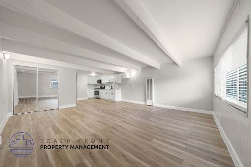 A spacious room with wooden flooring and white walls, with a logo for Beach Front Property Management in the bottom left corner.