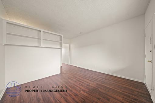 A room with wooden floors and white walls, with a logo for Beach Front Property Management in the bottom left corner.