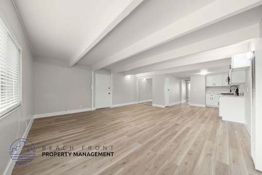 A spacious room with wooden flooring and white walls, with a logo for Beach Front Property Management in the bottom left corner.