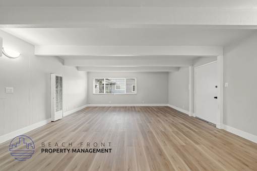 A spacious room with wooden flooring and white walls, with a logo for Beach Front Property Management in the bottom left corner.