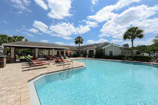 A swimming pool surrounded by a patio with chairs and a building in the background.