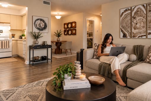 A woman is sitting on a couch using a laptop in a well-lit living room.