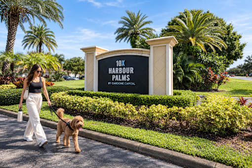 A woman walking her dog in front of a sign that says 10x Harbour Palms.