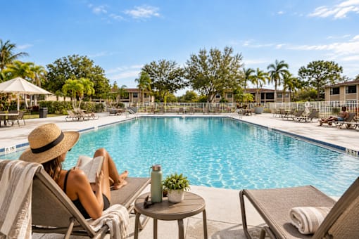 A woman in a straw hat is reading a book by the pool.