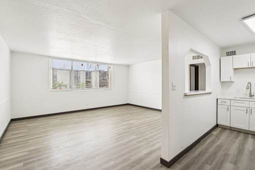 A room with white walls and wooden flooring, with a window and a small kitchenette.