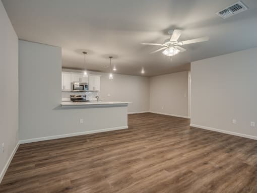 an empty living room with a ceiling fan and a kitchen in the background
