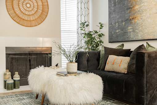 A living room with a fireplace, a white fluffy ottoman, a black couch, and a large painting on the wall.