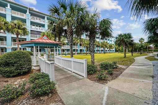 A gazebo sits in front of a building with a white picket fence and palm trees.